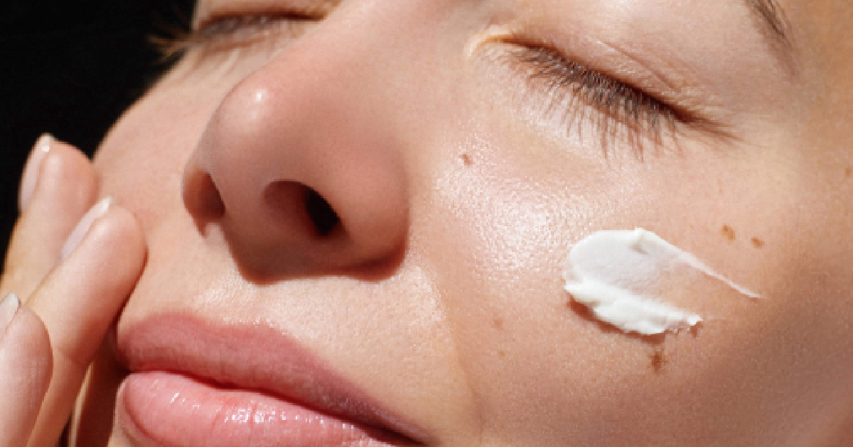 Close-up of a woman with glowing skin applying EltaMD® Skincare moisturizer to her cheek, eyes closed, showing smooth texture and a healthy complexion in Auburn, MA