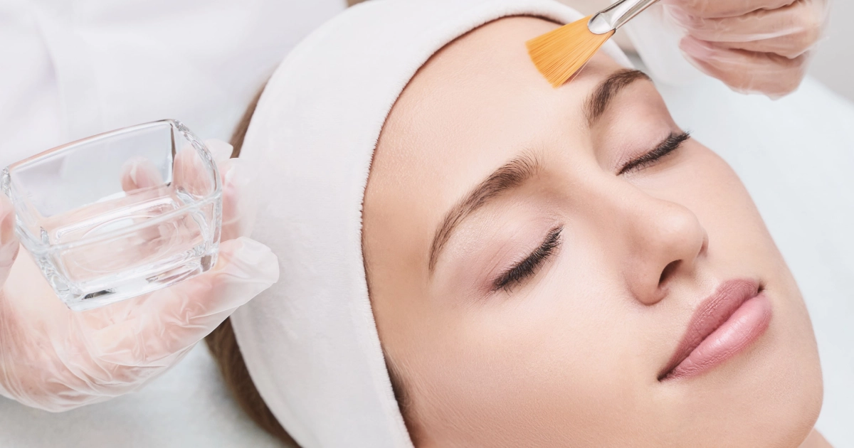 Woman receiving a chemical peel in a spa setting, eyes closed and relaxed, while a professional applies the solution to her forehead with a brush in Auburn, MA